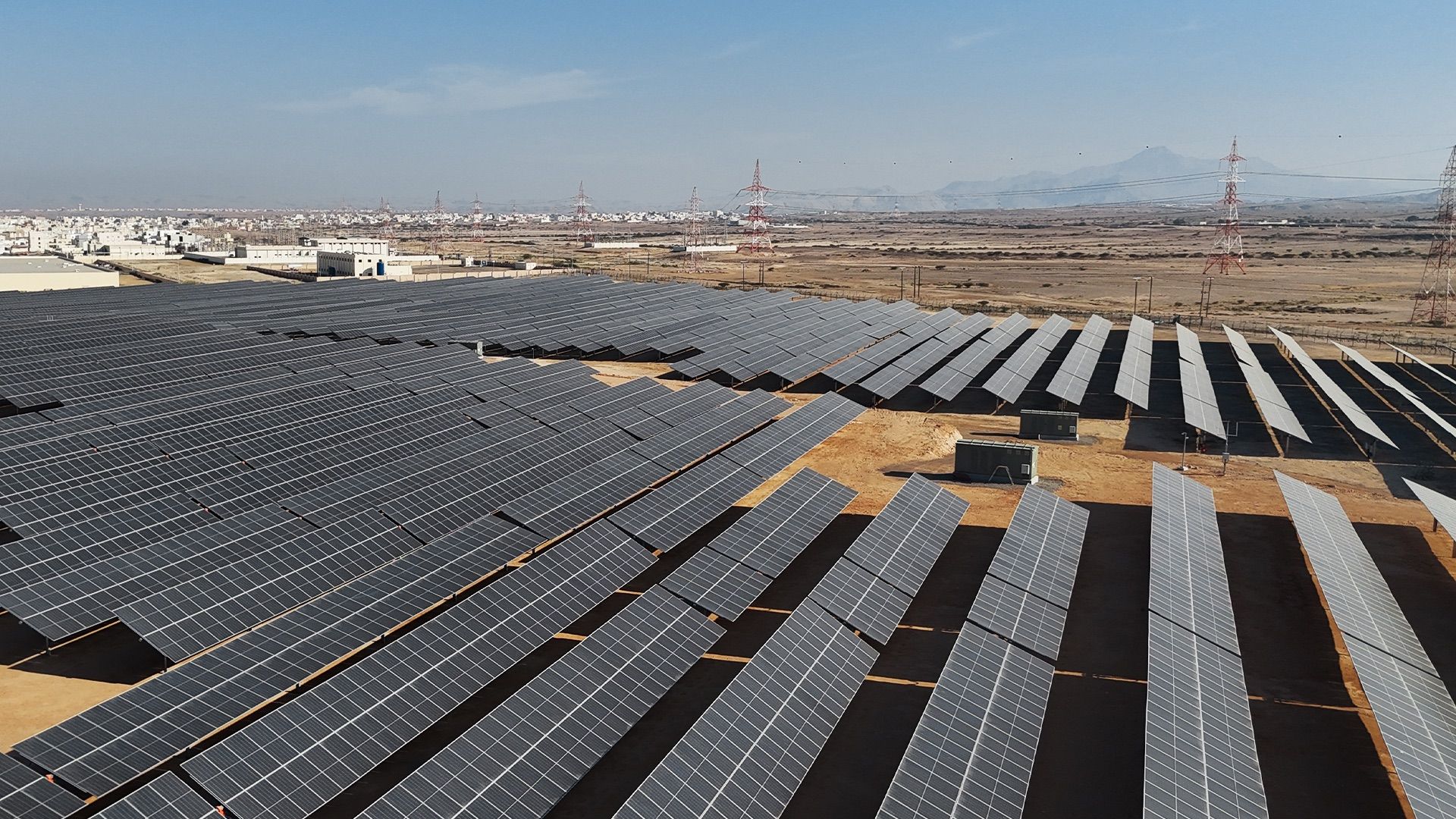 Rows of solar panels on the University of Buraimi campus with transmission pylons and mountains behind